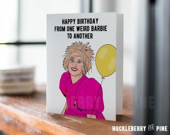 Pode incluir: Um cartão de felicitações dobrado com o texto "HAPPY BIRTHDAY FROM ONE WEIRD BARBIE TO ANOTHER" e uma ilustração de uma mulher com cabelo loiro e um vestido rosa segurando um balão amarelo. O cartão está em uma superfície de madeira.