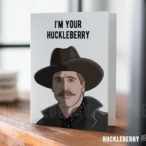 May include: A folded greeting card with the text "I'M YOUR HUCKLEBERRY" above a cartoon illustration of a man in a cowboy hat. The card has a white background and the words "HUCKLEBERRY AND PINE" are at the bottom.
