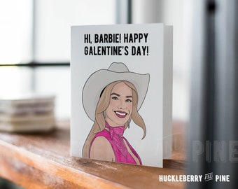 Pode incluir: Um cartão de felicitações dobrado com o texto "HI, BARBIE! HAPPY GALENTINE'S DAY!" apresenta uma ilustração de uma mulher usando um chapéu de cowboy e roupa rosa. O cartão está em uma superfície de madeira.