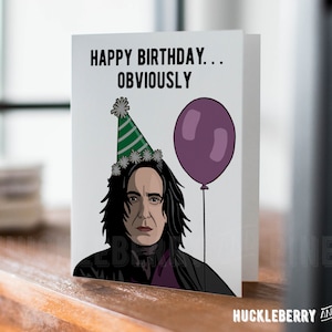 May include: A white greeting card with the text "HAPPY BIRTHDAY... OBVIOUSLY" and a cartoon illustration of a person wearing a party hat and holding a purple balloon. The card is on a wooden surface.