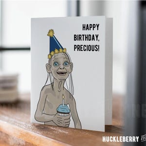 May include: A white greeting card featuring a cartoon illustration of Gollum from Lord of the Rings wearing a party hat and holding a cupcake with a lit candle. The text reads "HAPPY BIRTHDAY, PRECIOUS!" The card is on a wooden surface.