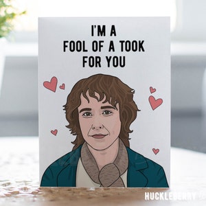 May include: A greeting card with the text "I'M A FOOL OF A TOOK FOR YOU" above an illustration of a person with brown hair and a scarf, surrounded by red hearts. The card has a white background.