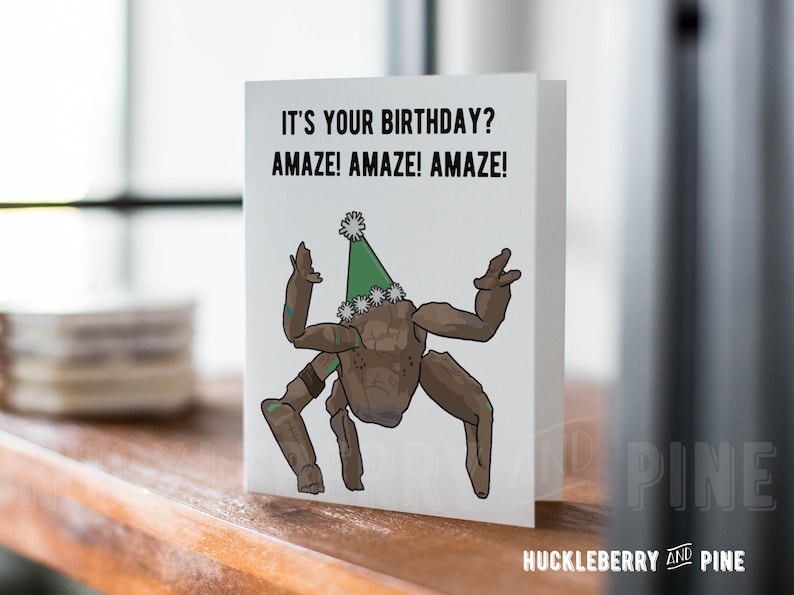 May include: A folded birthday card with the text "IT'S YOUR BIRTHDAY? AMAZE! AMAZE! AMAZE!" and a cartoon illustration of a brown creature wearing a green party hat. The card is white with the Huckleberry and Pine logo.