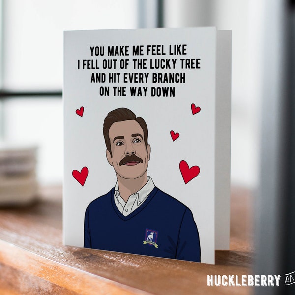 Op de afbeelding: Een wenskaart met een cartoon illustratie van een man met een snor en rode harten. De kaart toont de tekst: "YOU MAKE ME FEEL LIKE I FELL OUT OF THE LUCKY TREE AND HIT EVERY BRANCH ON THE WAY DOWN."