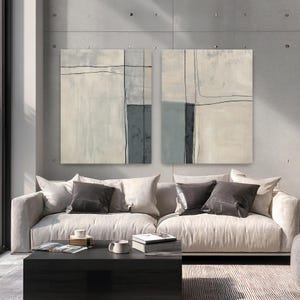 Peut inclure: Deux peintures abstraites au design minimaliste. Chaque toile pr&eacute;sente un fond clair avec des formes g&eacute;om&eacute;triques grises et noires. L'&oelig;uvre est accroch&eacute;e au-dessus d'un canap&eacute; clair avec des coussins gris fonc&eacute;.