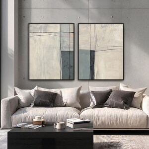 Peut inclure: Deux peintures abstraites encadr&eacute;es avec des bordures noires. L'&oelig;uvre pr&eacute;sente des tons sourds de beige, de gris et de bleu avec des accents lin&eacute;aires noirs. Les peintures sont expos&eacute;es au-dessus d'un canap&eacute; clair avec des coussins gris fonc&eacute;.
