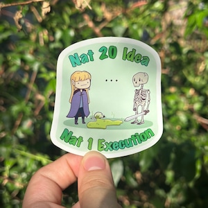 Puede incluir: Una pegatina con borde verde y texto "Nat 20 Idea" y "Nat 1 Execution". La pegatina presenta ilustraciones de dibujos animados de una persona con capa morada y un esqueleto con espada. También se representa una mancha verde y una calavera.