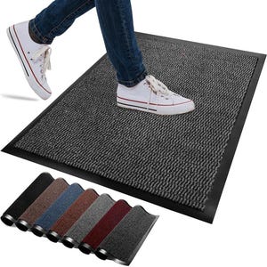 Puede incluir: Un felpudo rectangular gris con una superficie texturizada acanalada y un borde negro sólido. La alfombra se muestra en uso con zapatillas blancas y vaqueros azules, junto a una fila de seis alfombras adicionales en varios colores distintos.