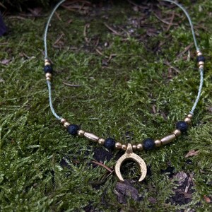 Op de afbeelding: Een gouden halve maan hanger ketting met zwarte lavastenen kralen op een lichtgroen koord.