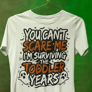May include: A white short-sleeve t-shirt featuring black and orange text that reads "YOU CAN'T SCARE ME I'M SURVIVING THE TODDLER YEARS" along with small graphic illustrations of bats, stars, and two ghosts on the front of the garment.