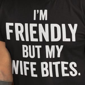 Peut inclure: Un t-shirt noir à manches courtes avec un texte blanc imprimé sur la poitrine qui dit I'M FRIENDLY BUT MY WIFE BITES. Le t-shirt présente une encolure ronde et une coupe standard, fabriqué dans un tissu noir uni avec des lettres grasses.