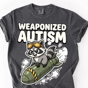 Może przedstawiać: Ciemnoszara koszulka z napisem "WEAPONIZED AUTISM" nad kreskówkowym szopem w okularach, jadącym na zielonym pocisku. Szop jest szary, biały i czarny. Pocisk ma żółte paski i białe smugi dymu.