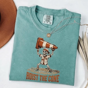 May include: Teal Comfort Colors t-shirt featuring a baseball-themed skeleton graphic. The skeleton is holding a traffic cone above its head. The text "HOIST THE CONE" is printed below the graphic. A silver necklace with a pendant is also visible.