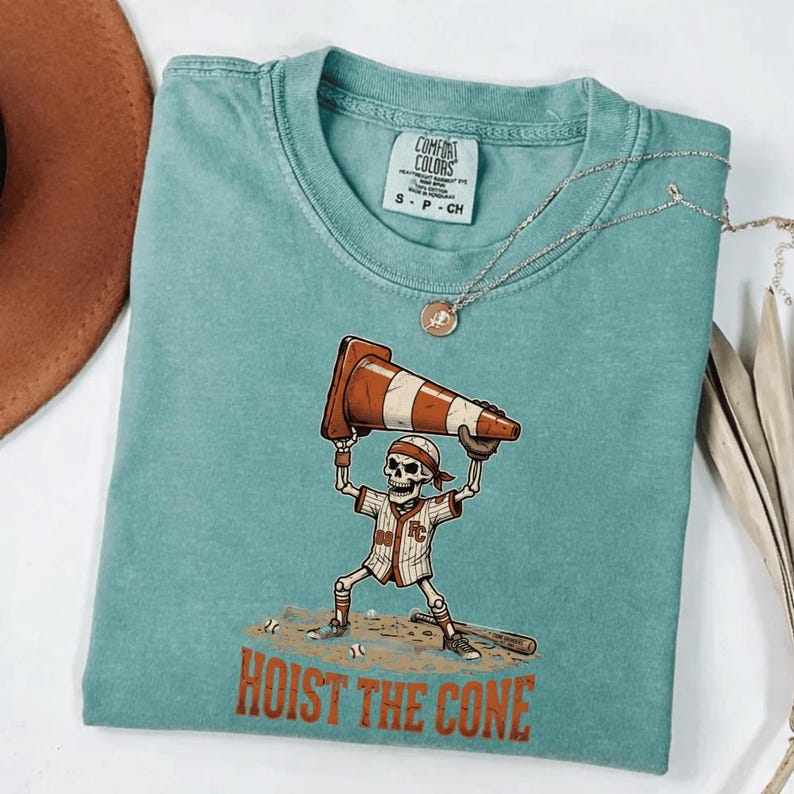 May include: Teal Comfort Colors t-shirt featuring a baseball-themed skeleton graphic. The skeleton is holding a traffic cone above its head. The text "HOIST THE CONE" is printed below the graphic. A silver necklace with a pendant is also visible.
