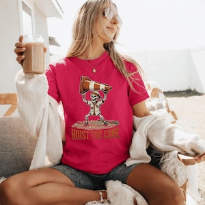 May include: A magenta t-shirt featuring a baseball-themed graphic. The design includes a skeleton holding a traffic cone above its head, with the text "HOIST THE CONE" below. The skeleton is wearing a baseball uniform.