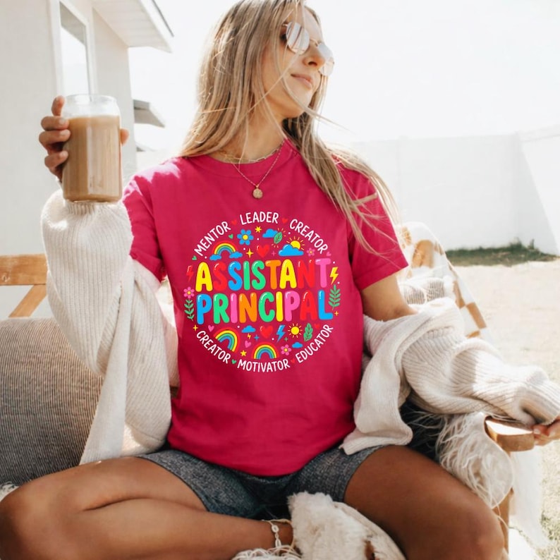 Peut inclure: Un t-shirt magenta avec l'inscription "Assistant Principal" dans un motif circulaire color&eacute;. Le motif comprend des arcs-en-ciel, des fleurs et les mots "Mentor, Leader, Creator, Motivator, Educator". Le t-shirt est &agrave; col rond.