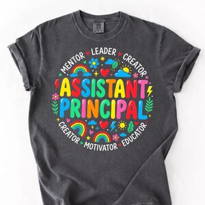 Peut inclure: T-shirt gris fonc&eacute; avec un graphisme color&eacute;. Le graphisme pr&eacute;sente les mots "ASSISTANT PRINCIPAL" en grandes lettres vives, entour&eacute;s de mots plus petits comme "Mentor" et "Educateur", ainsi que des arcs-en-ciel, des c&oelig;urs et des fleurs.