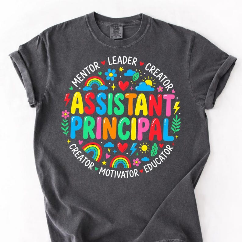 Peut inclure: T-shirt gris fonc&eacute; avec un graphisme color&eacute;. Le graphisme pr&eacute;sente les mots "ASSISTANT PRINCIPAL" en grandes lettres vives, entour&eacute;s de mots plus petits comme "Mentor" et "Educateur", ainsi que des arcs-en-ciel, des c&oelig;urs et des fleurs.