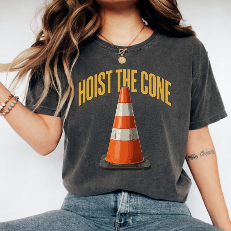 May include: Dark gray t-shirt with the text "HOIST THE CONE" in yellow above a cartoon traffic cone. The cone is orange and white, with a black base. The shirt is worn by a person.
