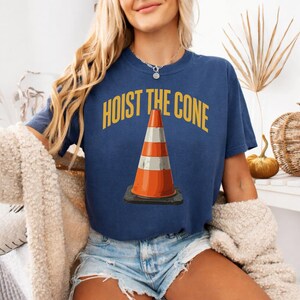 May include: Navy blue t-shirt with the text "HOIST THE CONE" in yellow above a graphic of an orange and white traffic cone. The cone is on a black base. The shirt is worn by a person.