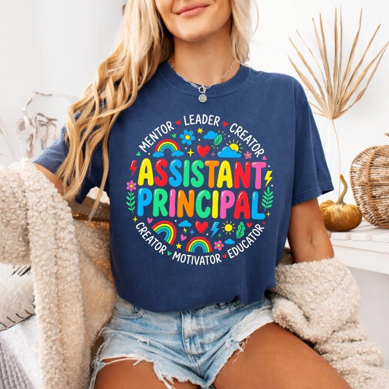 Peut inclure: Un t-shirt bleu marine avec un graphisme color&eacute; affichant "Assistant Principal" en grandes lettres multicolores. Le motif comprend des arcs-en-ciel, des c&oelig;urs et des &eacute;l&eacute;ments floraux, avec des mots comme "Mentor" et "Leader".