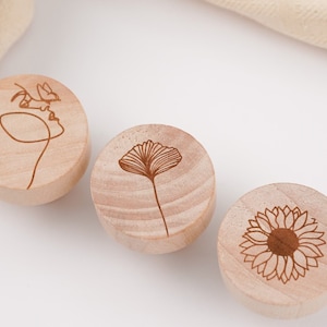 Könnte beinhalten: Drei runde Holz-Untersetzer mit verschiedenen gravierten Designs. Der erste Untersetzer zeigt eine Linienzeichnung eines Gesichts mit einem Schmetterling. Der zweite Untersetzer zeigt eine Linienzeichnung eines Ginkgoblattes. Der dritte Untersetzer zeigt eine Linienzeichnung einer Sonnenblume.