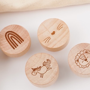Könnte beinhalten: Vier runde Flaschenverschlüsse aus Holz mit verschiedenen gravierten Designs: ein Regenbogen, ein Katzenkopf, ein Einhorn und ein Löwenkopf.