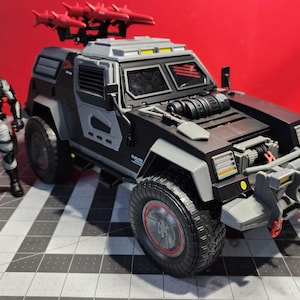 Puede incluir: Un vehículo militar de juguete negro y gris con un lanzamisiles rojo en la parte superior. El vehículo tiene un parachoques delantero grande con una rejilla de metal y un esquema de pintura negro y gris. El vehículo está sobre una superficie de rejilla gris y negra.