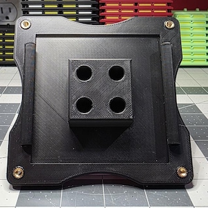 May include: Black 3D-printed stand with a square base and a central block featuring four circular holes. The stand has rounded edges and is secured with gold-colored screws. The textured surface adds a unique aesthetic.