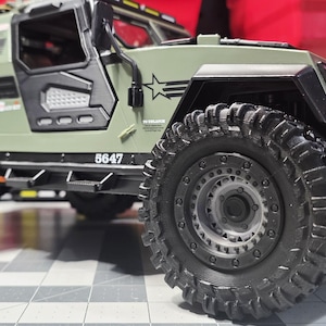 May include: An olive green toy military vehicle with large, black, off-road tires. The vehicle has a black front bumper with yellow accents and the number "6647" printed on the side. A star graphic is visible.