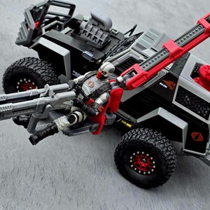 May include: A toy military vehicle in black, gray, and red, with large off-road tires and a mounted weapon. A figure in a white and gray uniform with a red helmet is seated in the vehicle. The vehicle has a crane arm.