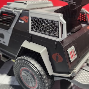 Puede incluir: Un vehículo militar de juguete negro y gris con una jaula gris en la parte trasera. El vehículo tiene un logotipo de Cobra en el lateral y luces rojas en la parte trasera.