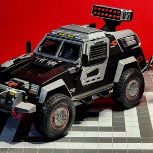 May include: A black and silver toy vehicle with a red light bar on top. The vehicle has a large front bumper with a metal grill and a silver side panel. The vehicle is on a gray and white checkered surface. A gray toy soldier stands next to the vehicle.