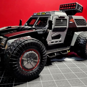 Op de afbeelding: Een zwarte speelgoedtruck met een grijs en zilveren carrosserie, rode lichten op het dak en grote zwarte banden. De truck heeft een gedetailleerd ontwerp met een grille, koplampen en een zijdeur.