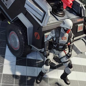 May include: A grey and black toy military vehicle with a red and black figure sitting on the back. The vehicle has the word "TURBO" printed on the side.