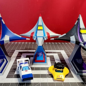 May include: Three blue, grey, and black toy spaceships with yellow accents on stands. The spaceships are arranged in a row on a grey and black chequered surface. There are two smaller toy vehicles in front of the spaceships.