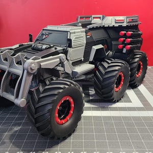 May include: A black and gray toy truck with large tires and a red and black design. The truck has a scoop on the front and a bed in the back. The truck is on a gray surface.