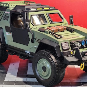 May include: A green and black toy military vehicle with a roll cage, a black door, and a black bumper with a yellow tow hook. The vehicle has a black grill with two blue lights on top. The vehicle has a black number plate that says "5647".