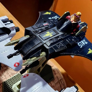 Puede incluir: Una nave espacial de juguete negra y dorada con una figura de piloto en la cabina. La nave espacial tiene una envergadura de unos 30 cm y presenta un símbolo de Cobra en el lateral. La nave espacial está hecha de plástico y tiene un diseño elegante y futurista.