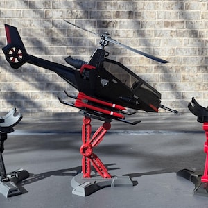 May include: A black model helicopter with red accents, mounted on a red and gray stand. The helicopter has a detailed design, including a rotor and missiles. Two additional stands are on either side.