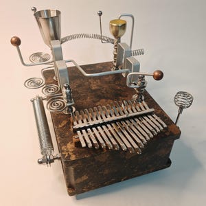 May include: A kalimba instrument featuring a rectangular wooden base with a mottled brown and black finish. The top is adorned with various silver-toned metal components, including coiled wire springs, a small funnel, a gold-colored cup, and metal rods.