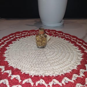 May include: A small glass bottle filled with gold flakes sits on a handmade, round crochet doily. The doily features a cream-colored center with a red and cream scalloped border. A white potted plant is in the background.