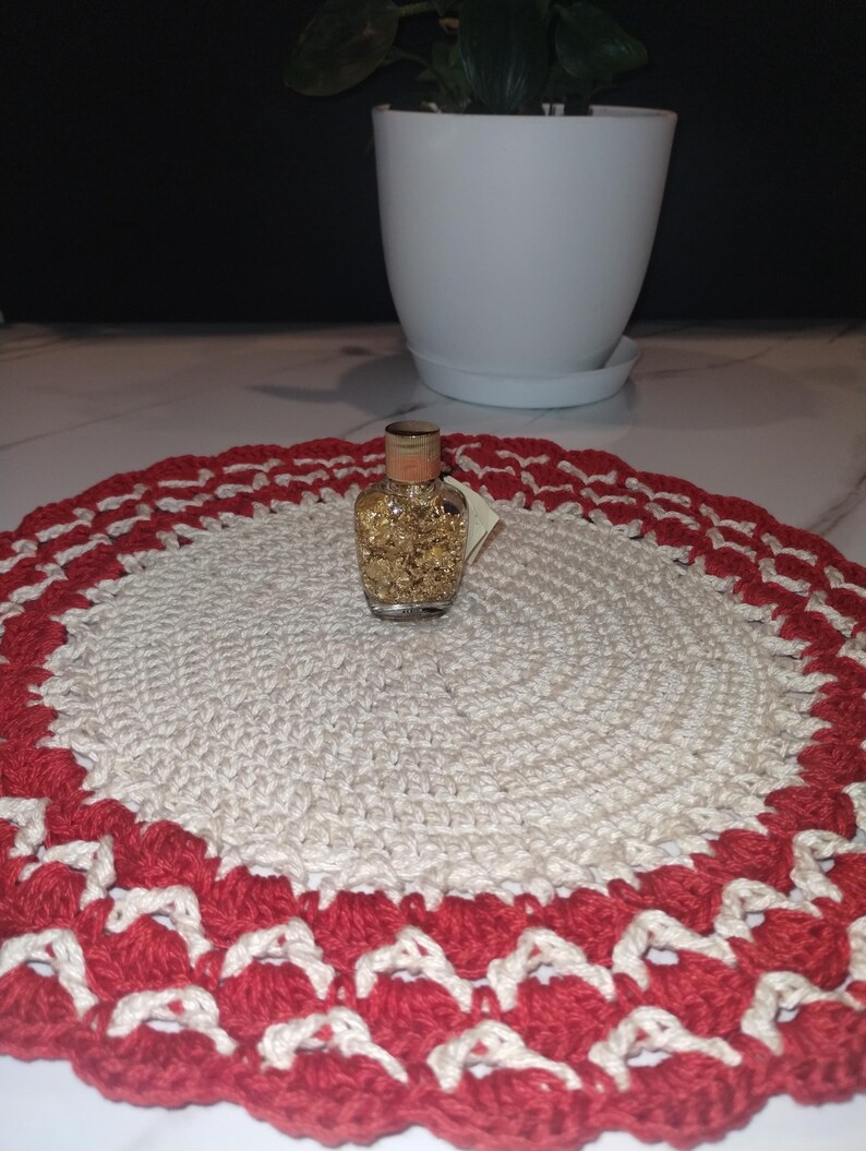 May include: A small glass bottle filled with gold flakes sits on a handmade, round crochet doily. The doily features a cream-colored center with a red and cream scalloped border. A white potted plant is in the background.