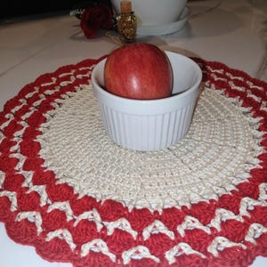 May include: A handmade, round crochet placemat with a scalloped red and cream border. A white bowl holds a red apple, centered on the placemat. The placemat is likely used for decorative purposes or to protect a table surface.