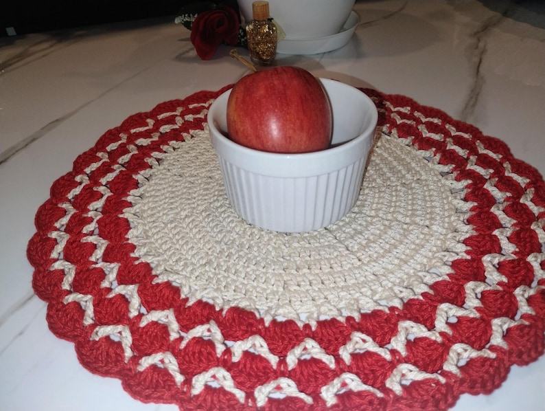 May include: A handmade, round crochet placemat with a scalloped red and cream border. A white bowl holds a red apple, centered on the placemat. The placemat is likely used for decorative purposes or to protect a table surface.