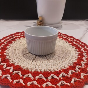 May include: A white ceramic ramekin sits on a handmade crochet doily. The doily features a cream-colored center with a scalloped red border. A white potted plant is in the background.