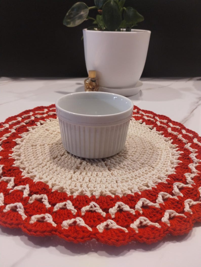 May include: A white ceramic ramekin sits on a handmade crochet doily. The doily features a cream-colored center with a scalloped red border. A white potted plant is in the background.