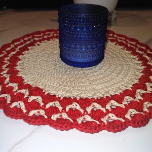 May include: A cobalt blue glass votive candle holder with a textured surface sits on a handmade, round crochet doily. The doily features a cream-colored center with a red scalloped border.