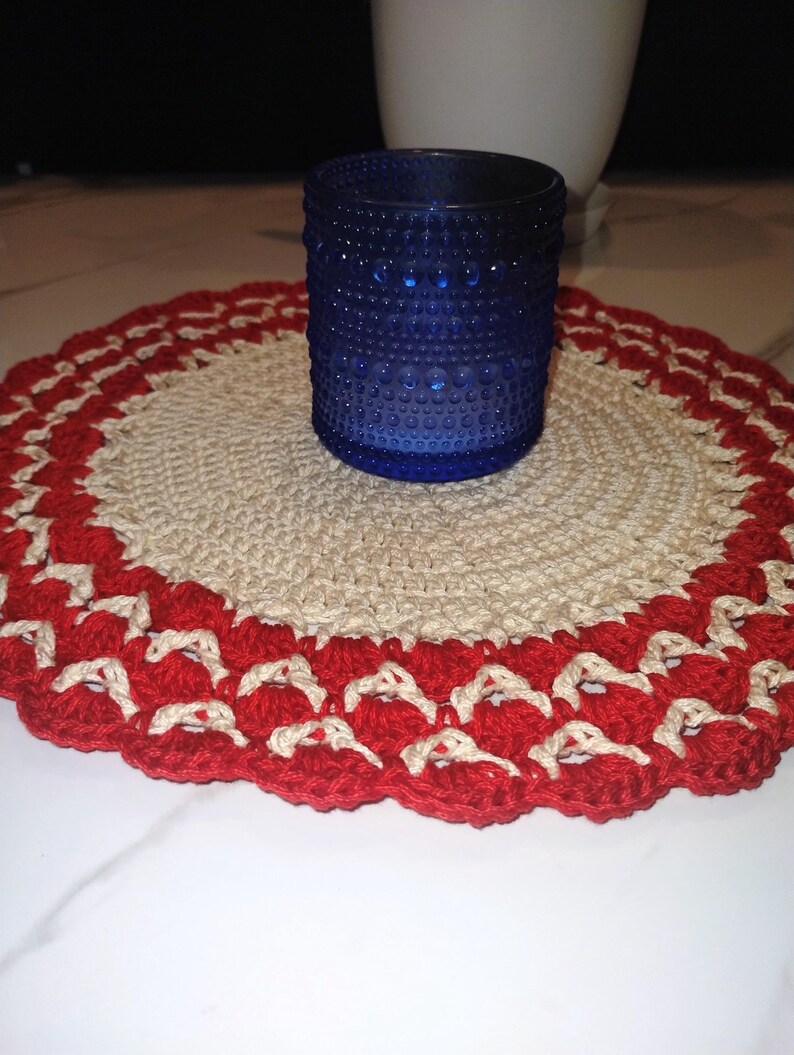 May include: A cobalt blue glass votive candle holder with a textured surface sits on a handmade, round crochet doily. The doily features a cream-colored center with a red scalloped border.