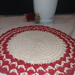 May include: A handmade, round crochet doily. The center is a natural beige color, surrounded by a scalloped border of red and cream-colored stitches. The doily is likely used for decorative purposes.